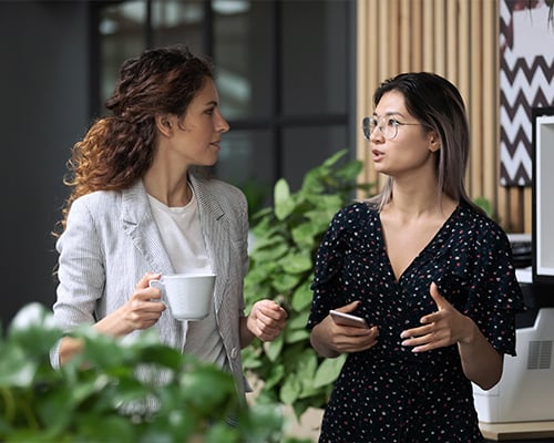 Diverse employees chatting during coffee break, walking in modern office, Asian businesswoman wearing glasses sharing ideas, discussing project with colleague, having pleasant conversation